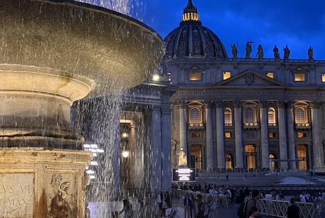 A nighttime photo of a fountain lit up in front of the Vatican.