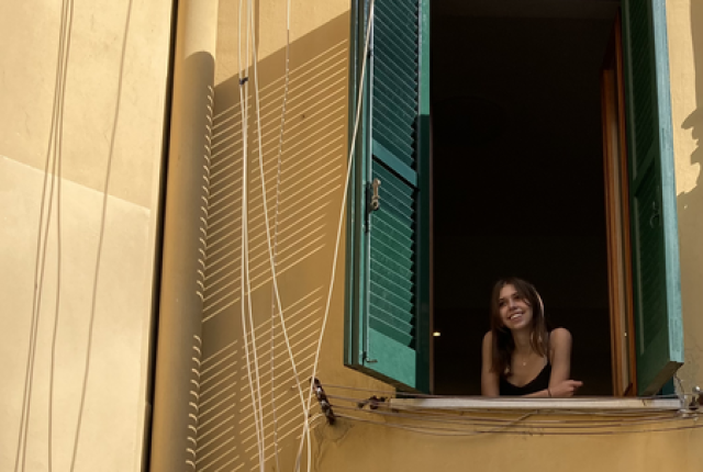A woman looks out of a window with green shutters against a yellow building.