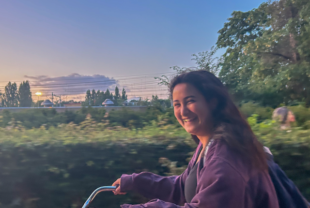 A student riding their bike and smiling with the sunsetting in the background.