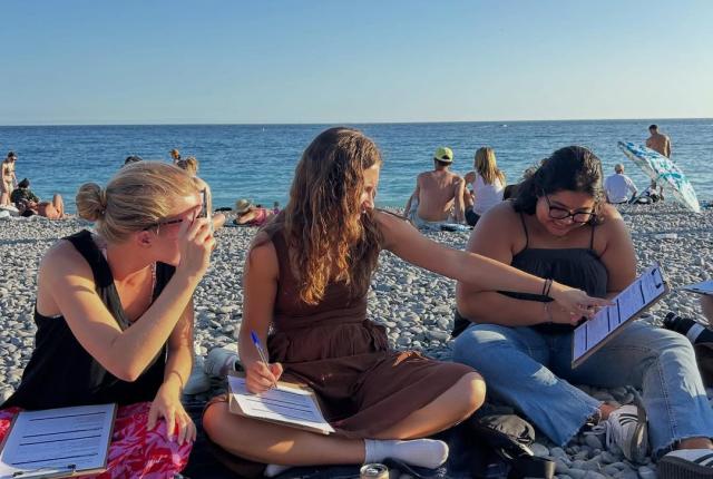 three students sitting on beach with clip boards