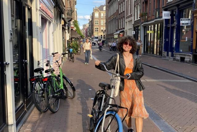 Student walking a bicycle along a brick-lined street in Amsterdam, with parked bikes, storefronts, and historic buildings stretching into the distance.
