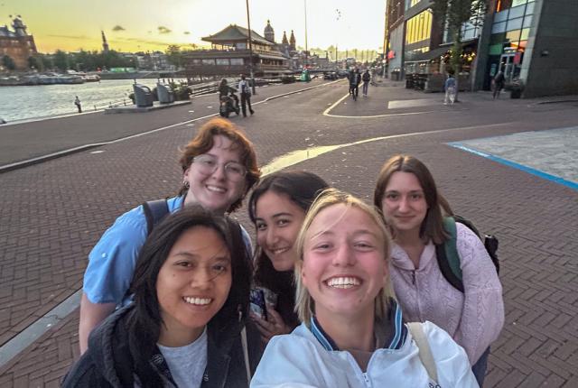 Five students taking a selfie along Amsterdam’s waterfront at sunset, with the water, historic buildings, and city skyline visible in the background.