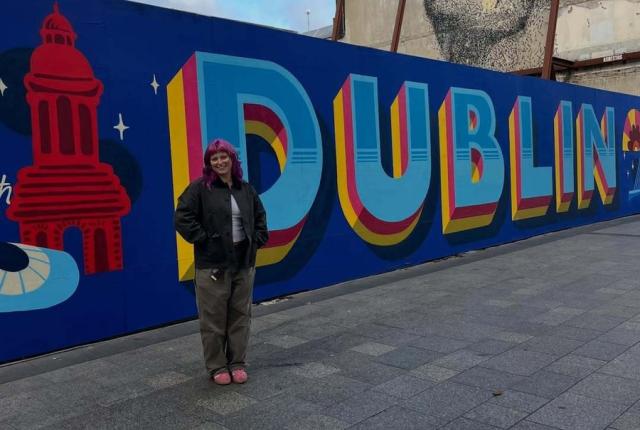 Student posing beside a large “Dublin” mural in the city center, with a historic building mural painted on a nearby wall.