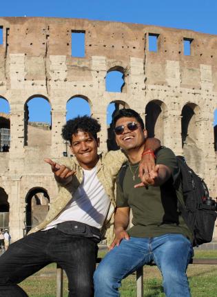 two male students pose for a fun photo in front of the Roman Colosseum in Italy