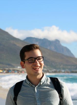 a male student smiling on the beach in Cape Town with moutains behind him