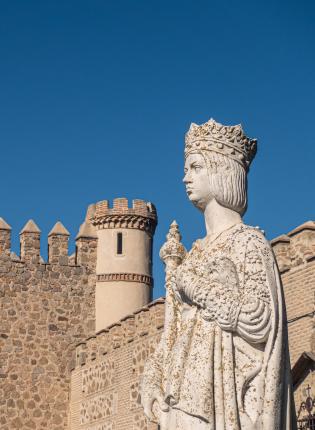 A white-washed statue of a person with a crown sits in front of a medieval building in Madrid, Spain.