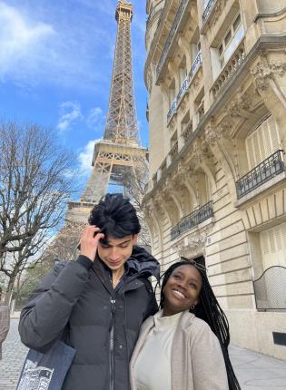 Two students, one taller than the other, are in front of the Eiffel Tower in Paris, France. One student smiles and the other is fixing his hair.