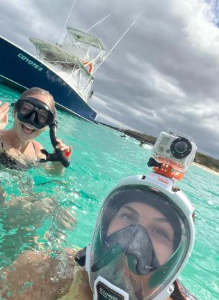 two students post for a photo in the ocean wearing snorkeling gear with a boat in the background