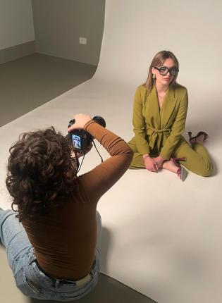 A fashion photoshoot with a model in a green pantsuit sitting on the floor, with the photographer sitting in front of them taking their photo