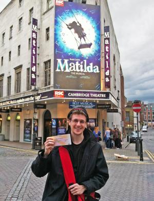 a male student standing outside of Cambridge Theatre with tickets to see the Matilda musical