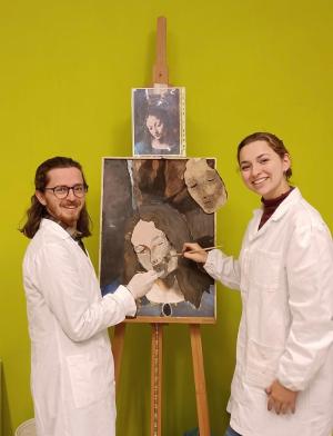 two students in white coats holding paint brushes in front of a painting on an easel