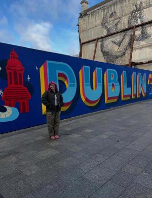 Student posing beside a large “Dublin” mural in the city center, with a historic building mural painted on a nearby wall.
