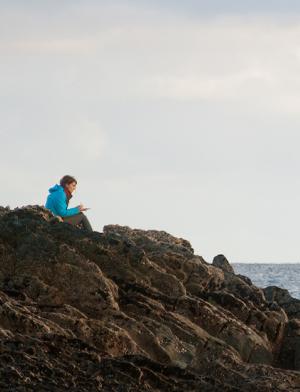 a student sitting on a rocky shore by the sea writing