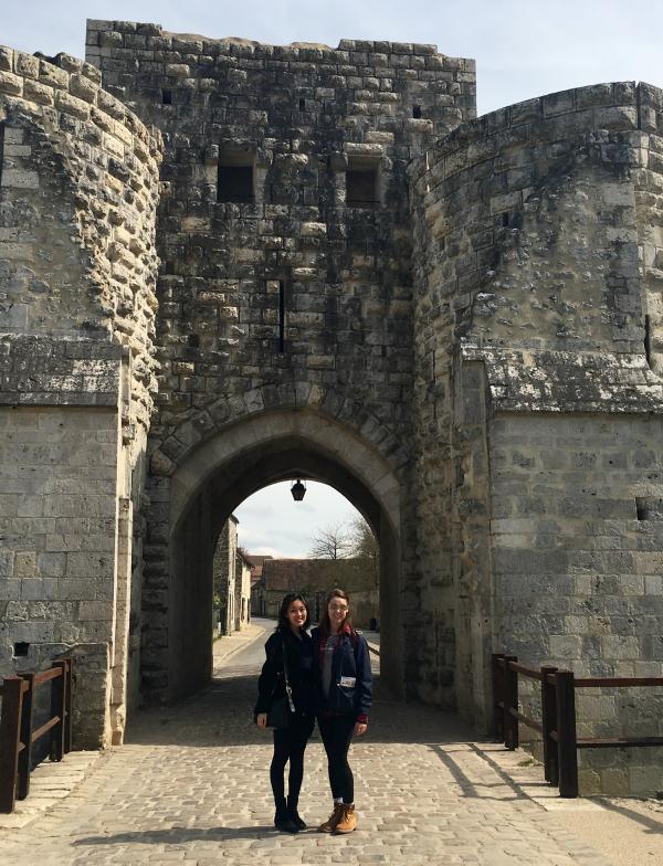 Two students stand in front of the gray stone walls of Provins. 