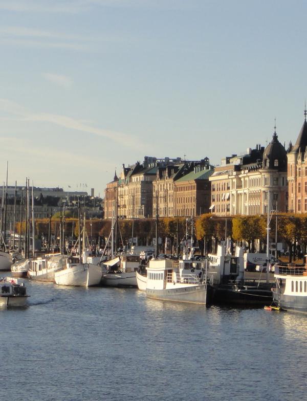 Stockholm Port on a sunny day. Numerous boats line the riverbank, and European-style buildings line the background.