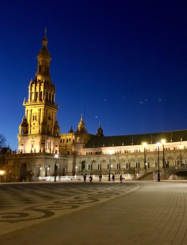 The Plaza de Espana photographed at night. A building with a tower and multiple spires sits in the background, with streetlights lighting up the square.
