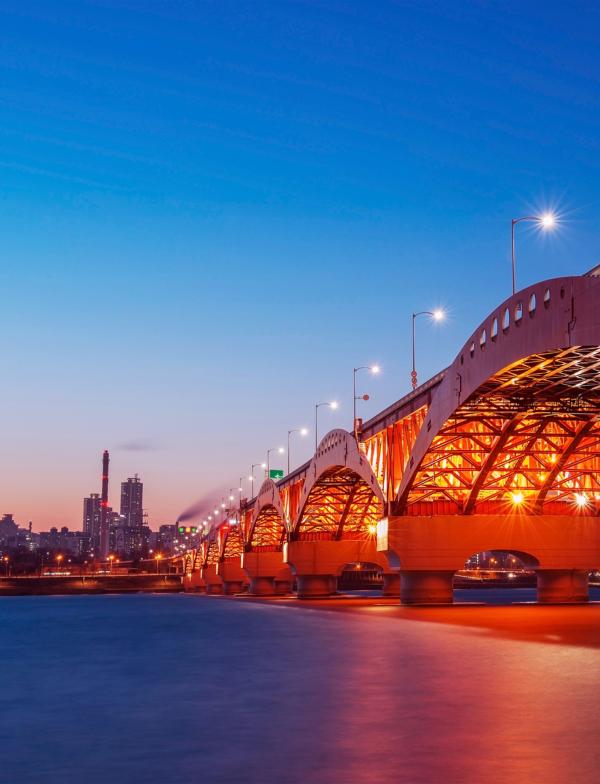 A large, steel, red underlit bridge crosses the Han River and leads to a city square.
