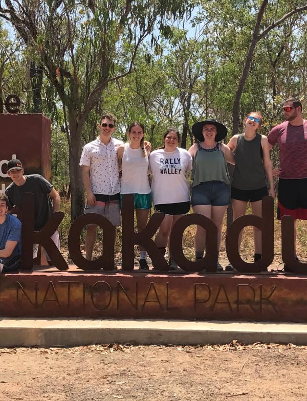 Students stand and sit around the entrance sign to Kakadu National Park. The sign reads "Welcome to the Aboriginal Land; Kakadu National Park."