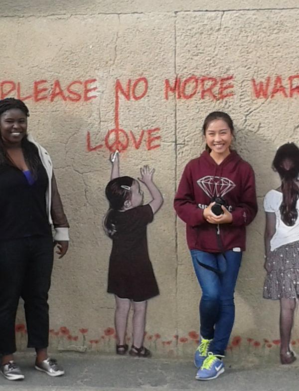 Students stand in front of a mural. The mural depicts two children writing "Please no more war, Love" with red chalk.