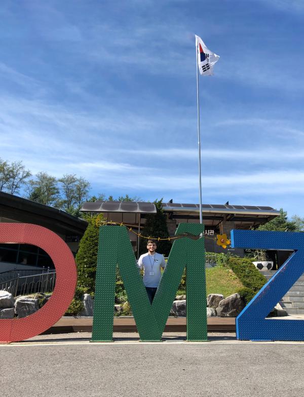 Large, colorful letters spell out "DMZ." A student stands behind the green M smiling at the camera.