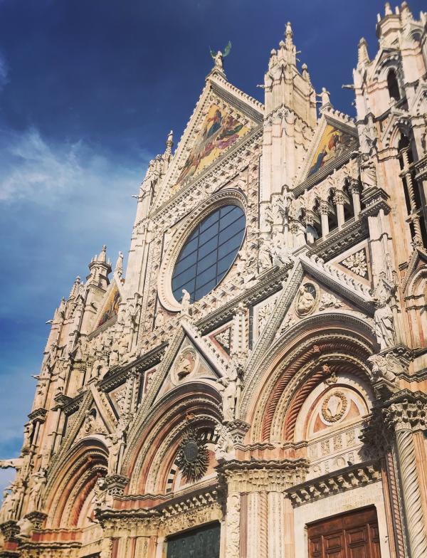 A cathedral in Siena, the Duomo Di Siena, on a sunny day. The exterior is an off-white color, with ornate details on the structure.