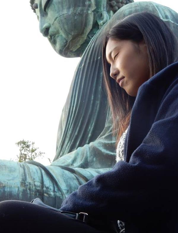 A perspective shot of a student rest in lotus pose while a much larger statue of Buddha in the same position sits in the background.