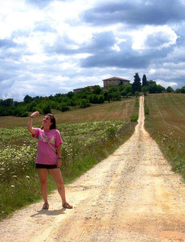 A student stands on a long trail path admiring a fruit in their hand. The path leads to a Tuscan-style home.