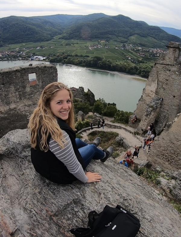 A student sitting atop Castle Ruins in Wachau. There is a river a few feet below, and hills and houses in the background landscape.