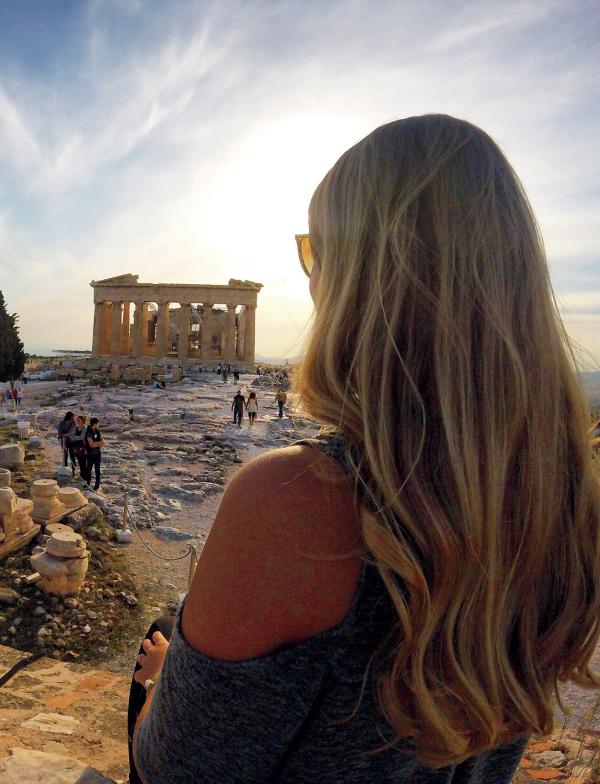 A student looks out over the ruins of the Parthenon in Athens. The sun is setting in the background.
