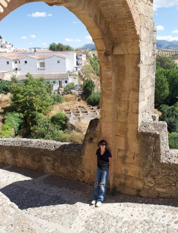 A student stands underneath a large tan arch in Setenil, Spain. The background includes trees and houses.