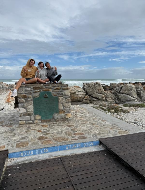 3 students sitting on a boulder in front of the ocean above a sign at the southern most point in africa