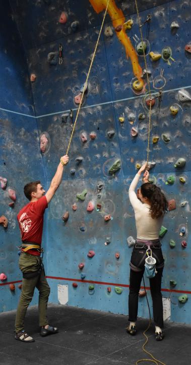 two students standing in front of a rock climbing wall holding on to rope and smiling