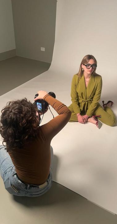 A fashion photoshoot with a model in a green pantsuit sitting on the floor, with the photographer sitting in front of them taking their photo
