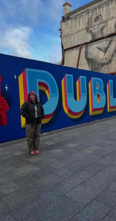 Student posing beside a large “Dublin” mural in the city center, with a historic building mural painted on a nearby wall.