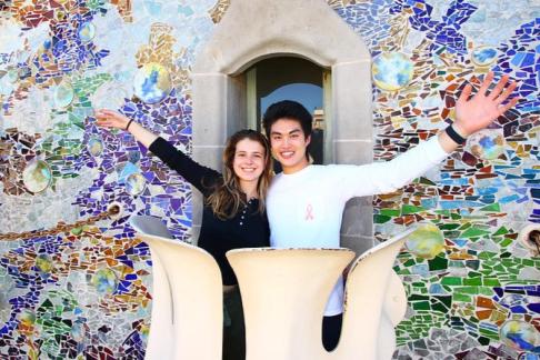 two students with arms stretched out standing on a balcony in front of a gaudi mural wall