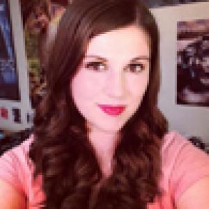 Smiling woman with long brown hair and pink top, colorful posters in background. This alt text was added with Al; accuracy may vary.