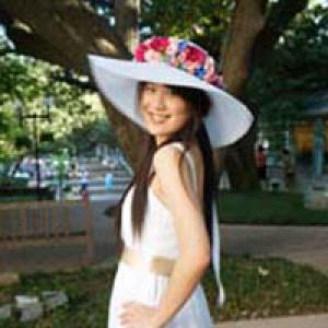 Smiling woman outdoors wearing a white hat with flowers under a large tree. This alt text was added with Al; accuracy may vary.