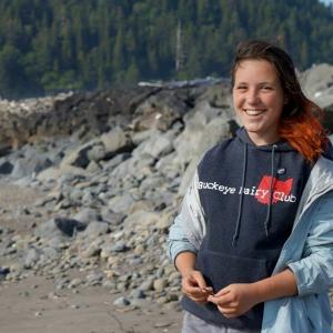 Smiling person wearing a hoodie on a rocky beach, with forested hills in the background. This alt text was added with Al; accuracy may vary.