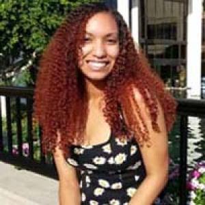 Smiling woman with curly hair, wearing a floral dress, outdoors by a railing. This alt text was added with Al; accuracy may vary.