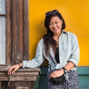 Smiling woman in denim by a yellow wall, holding a camera. This alt text was added with Al; accuracy may vary.