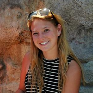 Young woman smiling against a rock wall, wearing sunglasses on head. This alt text was added with Al; accuracy may vary.
