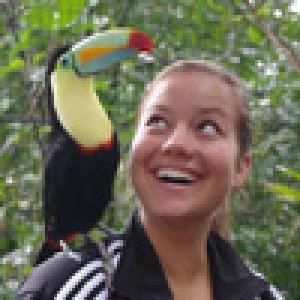 Smiling woman with a toucan on her shoulder in a forest setting. This alt text was added with Al; accuracy may vary.