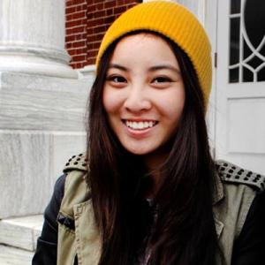 Smiling person with long hair, wearing a yellow beanie, sitting outside near columns. This alt text was added with Al; accuracy may vary.