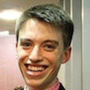 Smiling person in a suit indoors with short hair. This alt text was added with Al; accuracy may vary.