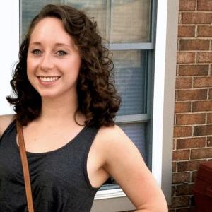 Smiling person with curly hair stands outdoors near a brick wall and window. This alt text was added with Al; accuracy may vary.