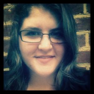 Smiling person with dark hair and glasses, brick wall background. This alt text was added with Al; accuracy may vary.