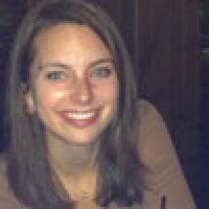 Smiling person with brown hair wearing a brown top indoors. This alt text was added with Al; accuracy may vary.