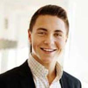 Smiling person with short hair in a well-lit room, wearing a suit and checkered shirt. This alt text was added with Al; accuracy may vary.