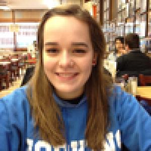 Smiling person in a blue sweatshirt sitting in a busy cafe. This alt text was added with Al; accuracy may vary.