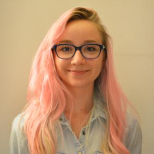 Young person with pink hair, glasses, and a denim shirt, smiling against a neutral background. This alt text was added with Al; accuracy may vary.